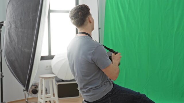 Man adjusting camera while seated in studio with green screen and softbox lighting; creative concentration.