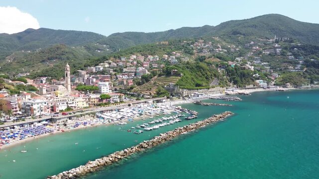 Moneglia, Liguria, Italy. Lateral aerial drone flight along Moneglia Beach and the small boat harbor, with Santa Croce Church, turquoise Ligurian Sea and green coastal hills.