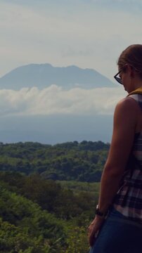 Vertical video. Young Female Traveler With Backpack Standing Above Tropical Forest And Watching Puncak Gunung Agung From Nusa Penida Under Dramatic Clouds, Adventure Travel And Exploration