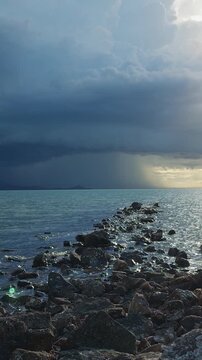 Vertical video. Slow Motion Wide Angle View of a Rocky Breakwater Leading Into a Tropical Sea Beneath Dramatic Storm Clouds, Sun Rays, Rainfall, Powerful Weather and Coastal Nature