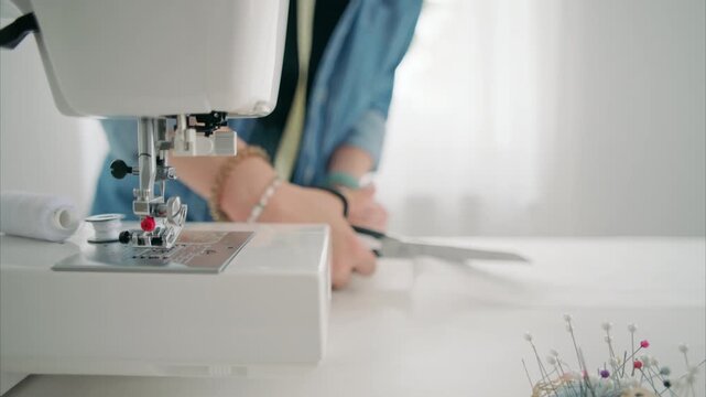 Woman using metal scissors to cut white cloth on desk with sewing machine and tailoring tools