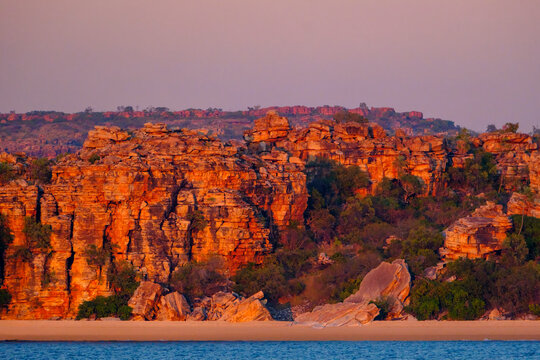 Sunrise lights up the red rust cliffs along the water in Kimberley, highlighting natural rock formations and vegetation