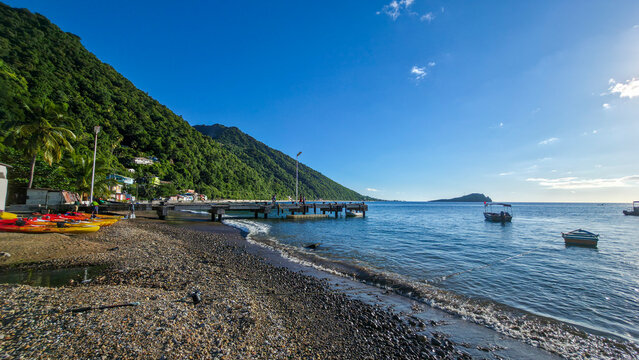 Kayaks rest on the shore while boats float in the water by the pier in Soufriere beach