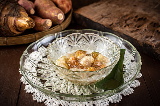 Homemade Thai Bua Loi Kaew dessert. Translucent orange sweet potato and taro tapioca balls in warm coconut milk soup. Served in glass bowls on a rustic wooden table with raw tubers. 