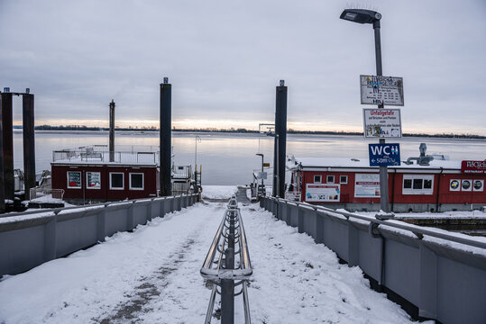 Snowy pathway leads to Elbe River ferry pier with view of cloudy winter horizon in Blankenese, Hamburg, Germany
