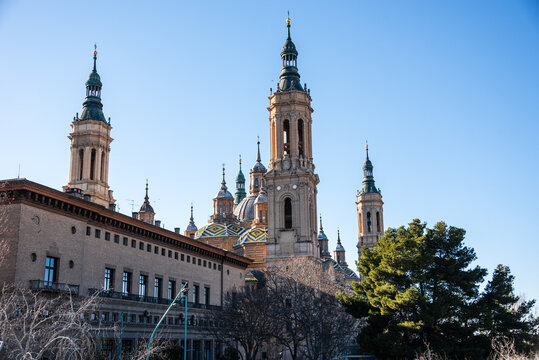 Tall towers of Baslica del Pilar rise above buildings and trees against a clear blue sky in winter in Zaragoza, Spain