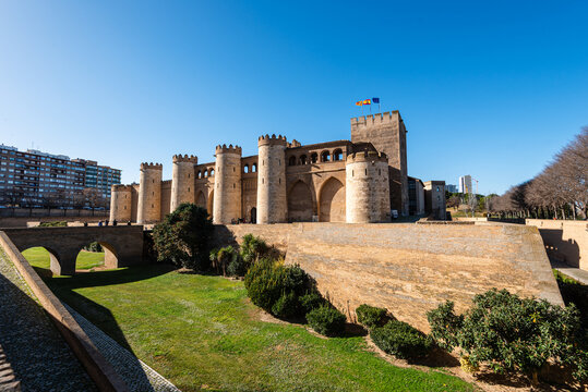 Panoramic view with restored outer walls and towers of Aljafera Palace in landscaped gardens under blue sky in Zaragoza