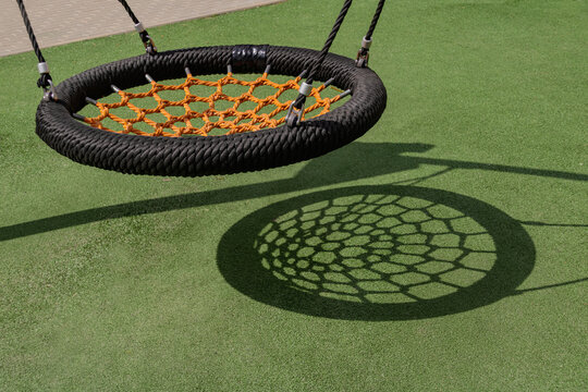 Round nest swing on outdoor playground casting geometric shadow on green turf, empty play equipment and urban recreation concept with copy space