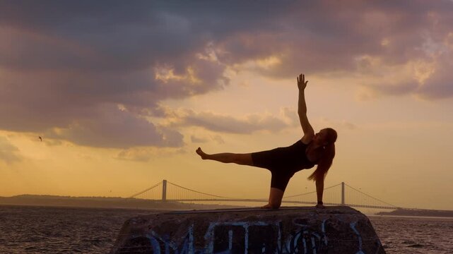 Woman executing side leg lift yoga posture in New York. Silhouette balancing on one knee with raised limb, Verrazzano Narrows Bridge, cloudy sunset sky, ocean.