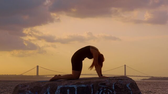 Woman practicing yoga cat pose at sunset in New York. Female silhouette doing back arch on stone ledge, Verrazzano Narrows Bridge, golden sky, water.