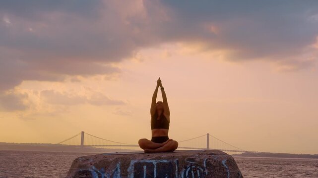 Woman meditating in lotus yoga position at sunset in New York. Front view of seated practitioner on concrete structure facing water with bridge skyline.