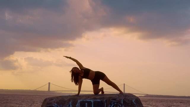 Woman performing kneeling side stretch yoga pose at sunset in New York. Full length body bending sideways on stone pier overlooking Verrazzano Narrows Bridge.