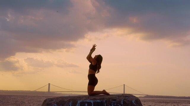Woman doing kneeling backbend yoga exercise at sunset in New York. Side profile with wrapped arms on waterfront block against Verrazzano Narrows Bridge sky.