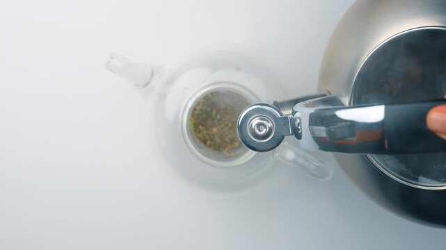 Pouring hot water from kettle into glass teapot. Top view, hand holding metallic kettle spout pouring clear liquid over green melissa leaves, white surface.