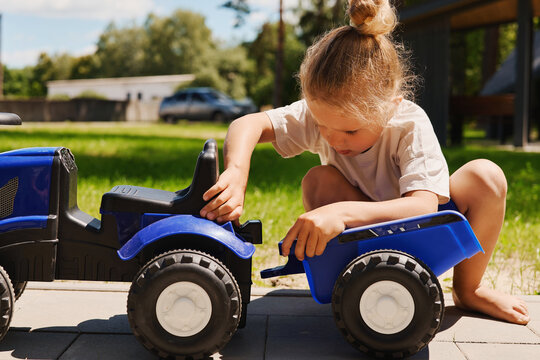 A young child sits on the ground, focused on connecting two toy trucks in a backyard. Trees and a car are visible in the background on a sunny day