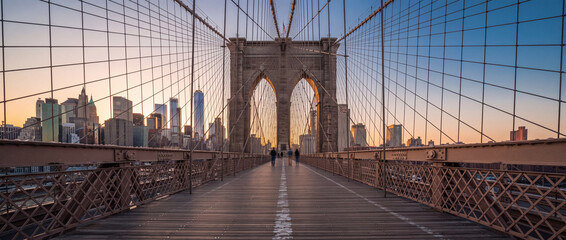 Fototapeta premium Brooklyn Bridge pedestrian walkway at sunrise with suspension cables framing the Lower Manhattan skyline in warm morning light, ideal for urban travel and tourism concepts