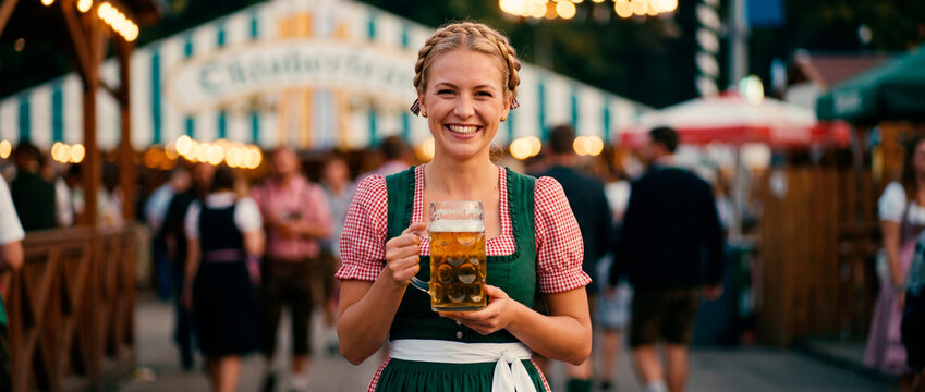 Smiling woman in traditional dirndl holding beer mug at Oktoberfest festival. Bavarian culture and german folk celebration concept. Evening beer garden party atmosphere.