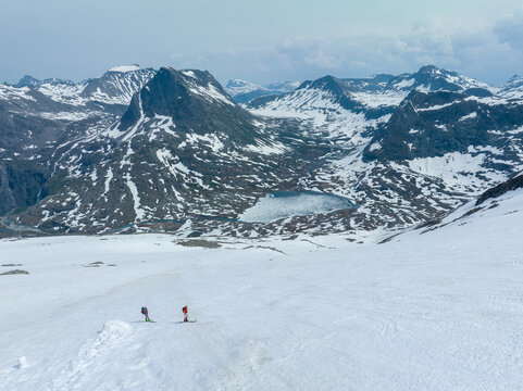 Aerial view of two skiers traversing a vast snow-covered slope near Alnestinden with a glacial lake and rugged mountain peaks in the background in Fjord, More og Romsdal, Norway.