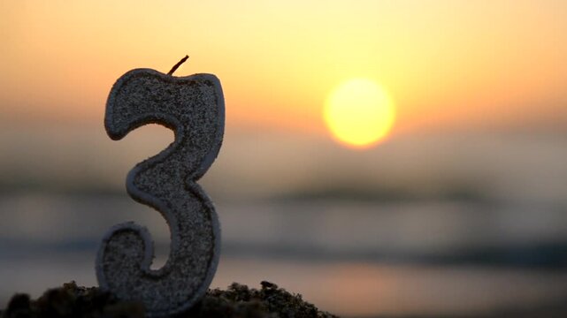 Candle in the shape of the number three 3 burning against background of blue sea waves, sun and orange sky in the sand on the beach of seashore at sunset and dawn. Concept Happy Birthday Holiday party