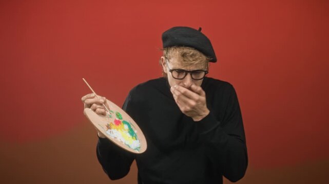Blond man painter holds wooden palette and brush, hand covering mouth gesture in studio with red wall; nausea hesitation.