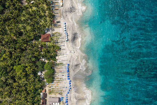 Aerial view of a tropical beach in Bali with lush palm trees, rows of traditional fishing boats lined up on the sand, and clear turquoise ocean water in Kuta Selatan, Bali, Indonesia.