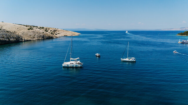 Aerial view of sailboats anchored in the clear blue waters of the Adriatic Sea near the rocky coastline of the island in Rab, Primorje-Gorski Kotar, Croatia.
