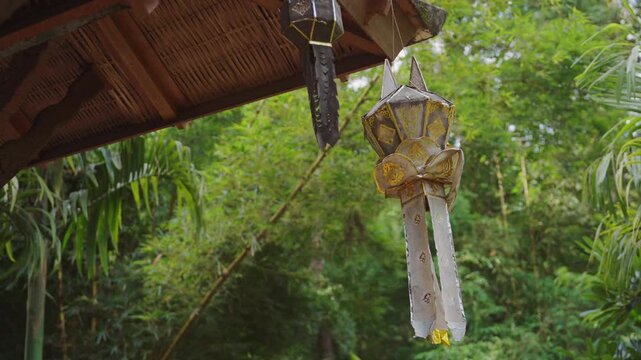 Traditional hanging lantern beneath wooden pavilion surrounded by tropical garden, Decorative Asian roof ornament with lush bamboo forest and green foliage outdoors 4k