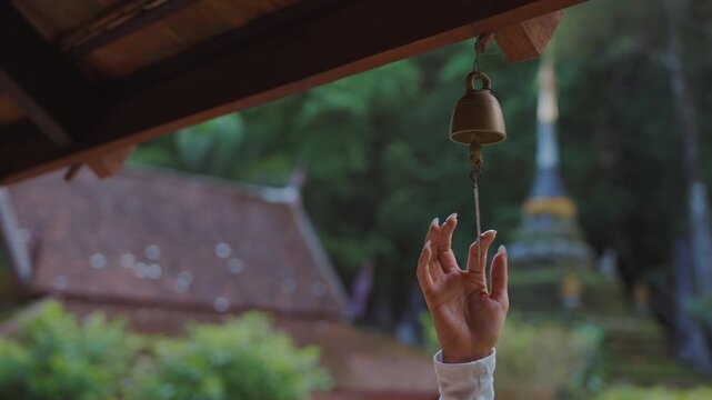 Woman touching hanging temple bell beneath traditional wooden pavilion, Buddhist prayer bell with leaf ornament at historic temple in Thailand 4k