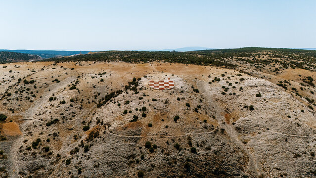 Aerial view of the checkered circular helipad on the rocky hill of Rab, surrounded by dry Mediterranean landscape and sparse vegetation in Rab, Primorje-Gorski Kotar, Croatia.