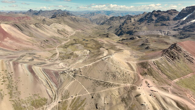 Aerial view of Vinicunca, the Rainbow Mountain, showcasing its colorful mineral layers, rugged Andean terrain, and winding trails under a blue sky in Quispicanchi Province, Cusco, Peru.