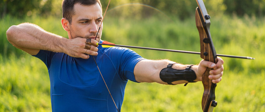 Archer aiming with recurve bow outdoors, pulling arrow string with focused concentration on sunny green field, perfect for sports training and active lifestyle themes