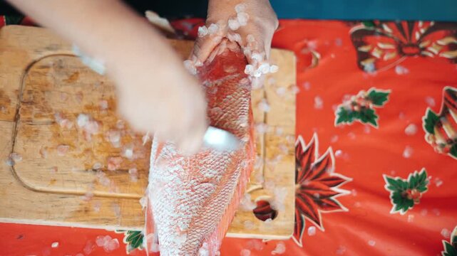 Hands using a knife to scale a fresh red snapper on a wooden board over a christmas tablecloth