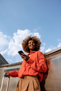 Woman using smartphone beside suitcase under blue sky