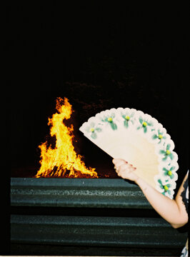 Hand holding traditional fan cooling a bonfire at night