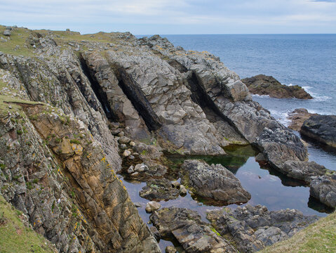 Tilted layers of ancient rock form dramatic coastal cliffs and tide pools at Bu Ness on Fair Isle Shetland UK beneath a pale blue sky