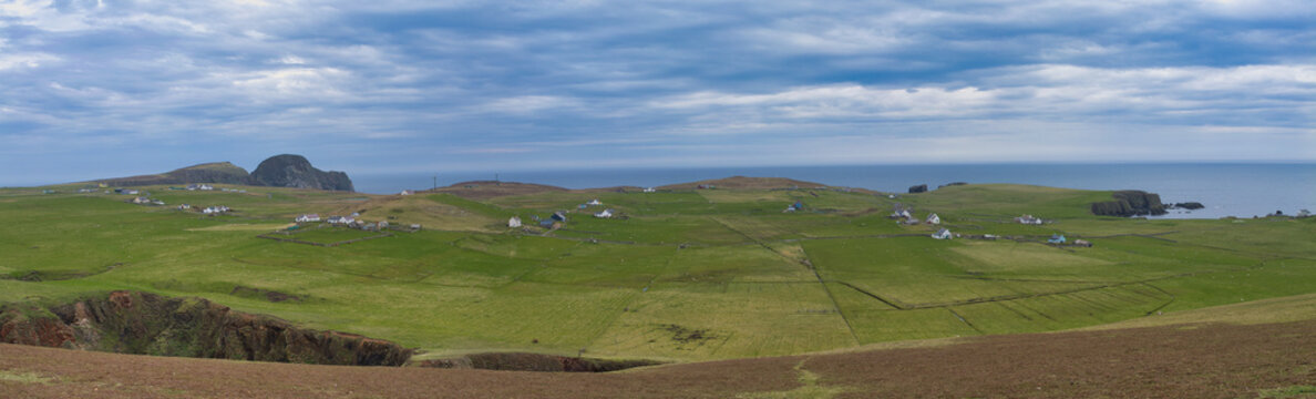 Scattered croft houses across green fields on south Fair Isle UK beneath a dramatic cloud filled sky and blue sea