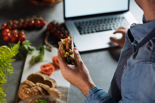 Man multitasking eating sandwich while working on laptop