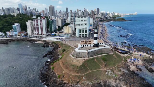 Cinematic aerial view of the historic Barra Lighthouse (Farol da Barra) and fort on the beautiful coastline of Salvador, Brazil.