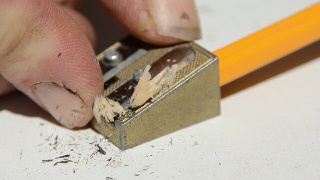 Pencil sharpening close up hand rotates graphite pencil in manual sharpener creating wood shavings in 4K Ultra HD footage.
