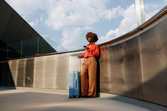 Woman checks smartphone beside suitcase outside building