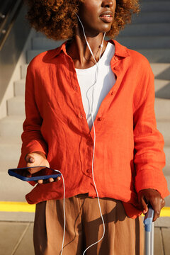 Woman with earphones holds smartphone beside suitcase