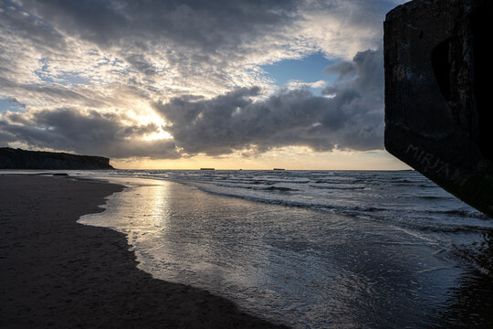 A concrete Mulberry harbour caisson remnant on the sandy beach of Arromanches-les-Bains in Normandy during a dramatic sunset with sun rays breaking through clouds.