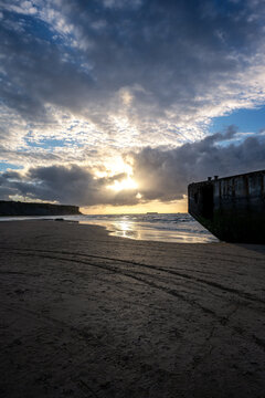 A concrete Mulberry harbour caisson remnant on the sandy beach of Arromanches-les-Bains in Normandy during a dramatic sunset with sun rays breaking through clouds.
