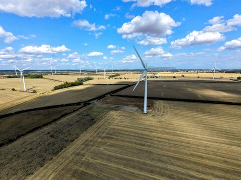 Aerial view of Burton Wold wind farm featuring turbine 1 amidst golden agricultural fields under a bright blue sky with scattered clouds in Burton Latimer, England, United Kingdom.