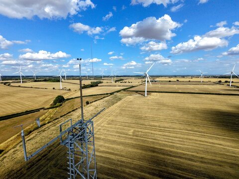 Aerial view of Burton Wold wind farm featuring multiple wind turbines set within vast golden agricultural fields under a bright blue sky in Burton Latimer, England, United Kingdom.