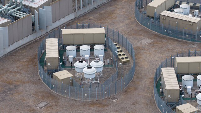 Aerial view of an industrial facility featuring circular fenced enclosures with storage tanks, cooling units, and piping infrastructure in Faversham, England, United Kingdom.