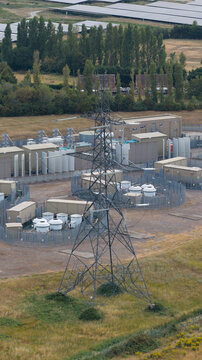 Aerial view of an electrical substation featuring a large metal pylon, industrial equipment, and transformers within a fenced enclosure in Faversham, England, United Kingdom.