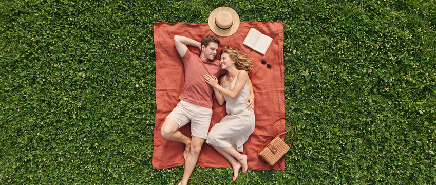 Romantic couple lying on picnic blanket outdoors on green clover lawn, top down view of young man and woman relaxing together during summer date, perfect for leisure travel and lifestyle