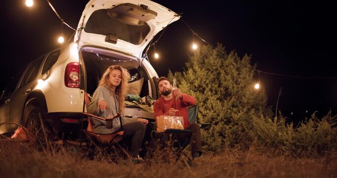 Friends enjoy a picnic dinner at night by the car under string lights in a field with trees around them