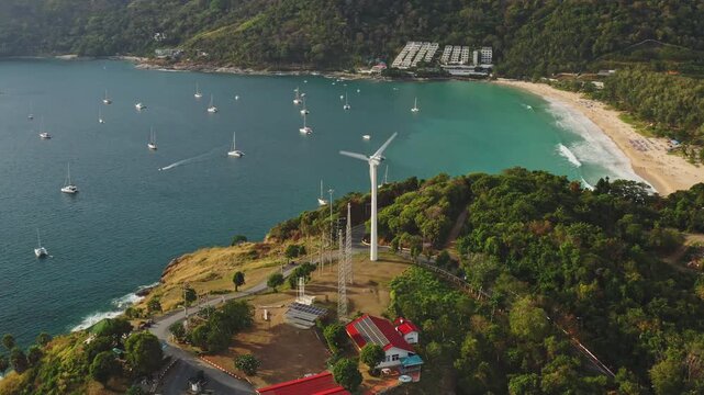 Windmill Viewpoint discovering Nai Harn beach with turquoise water, anchored yachts, and a wind turbine generating clean energy on the green tropical coastline of Phuket, Thailand. Aerial drone view